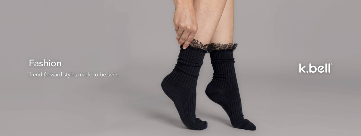A fashion promotional banner with the text 'Fashion' and the 'k.bell' logo. A woman wears lace-trimmed, ribbed black K.Bell socks on a warm gray background.
