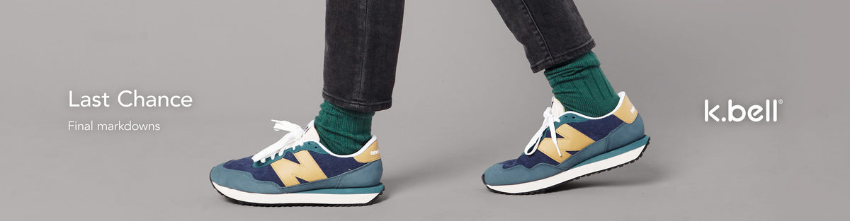 A person wearing dark jeans, green socks, and blue New Balance sneakers with yellow accents, shown from the knees down against a light gray background. Text on the left reads 'Last Chance – Final markdowns' and the 'k.bell' logo