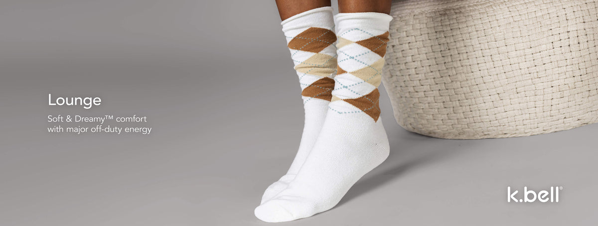 A person wearing white argyle socks with tan and beige diamond patterns, shown next to a woven basket on a light gray studio background. Text on the right reads 'Lounge – Soft & Dreamy™ comfort with major off-duty energy' and the 'k.bell' logo