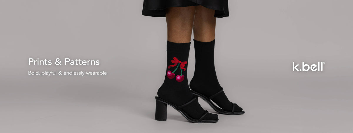 A model wearing black socks with a red cherry graphic, paired with black heeled sandals, against a light gray background with text reading 'Prints & Patterns – Bold, playful and endlessly wearable' and the 'k.bell' brand logo