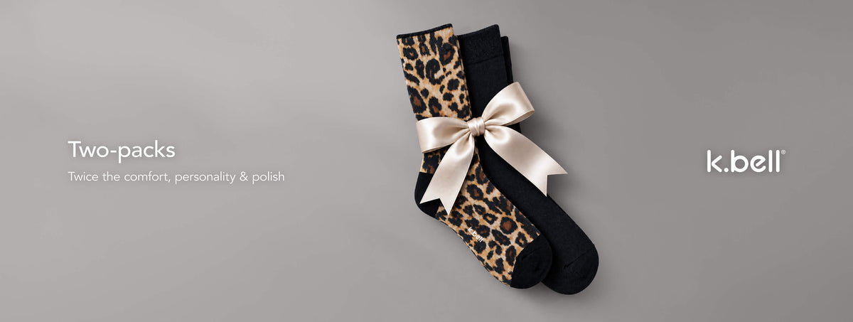 Two pairs of socks in a two-pack, featuring one pair in black and one pair in a leopard print pattern, tied together with a cream satin ribbon. The image is set against a solid gray background with text on the left reading 'Two-packs' and 'Twice the comfort, personality & polish', and the brand name 'k.