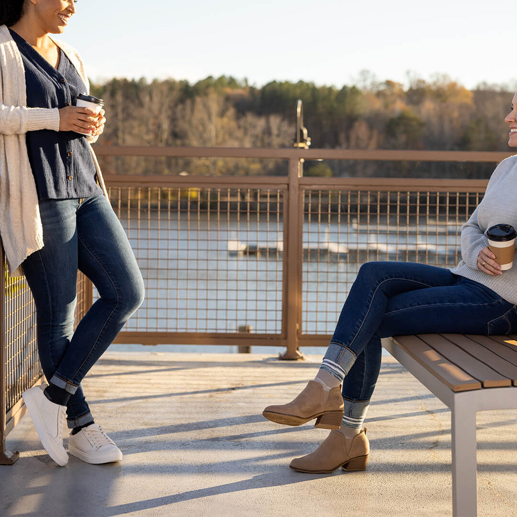 Two women on a wooden deck by a lake, one standing and one sitting on a bench, both holding coffee cups.
