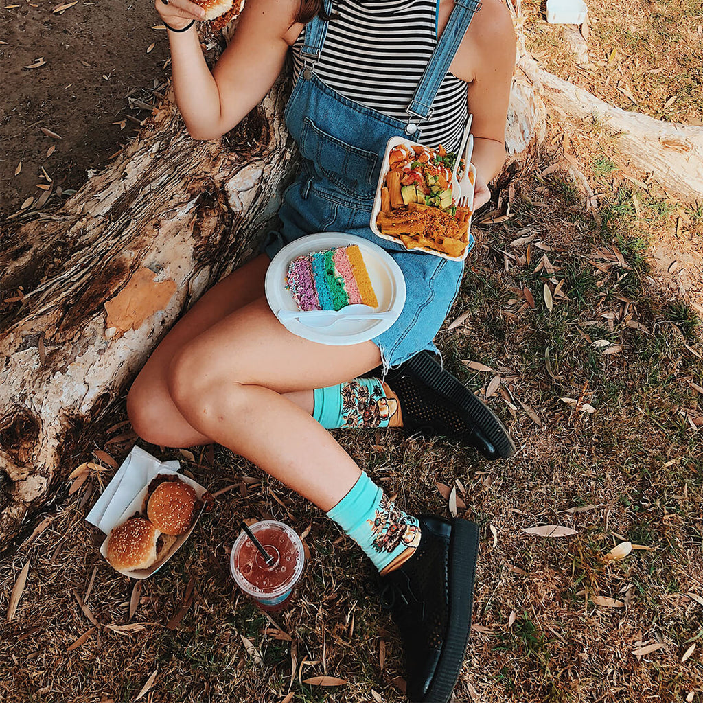 Person sitting outdoors with food, wearing denim overalls and colorful socks.