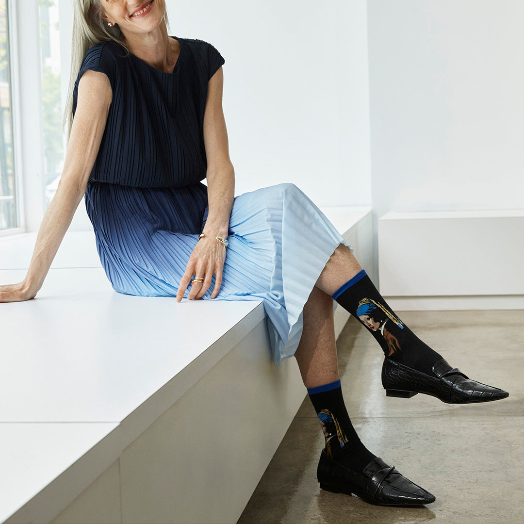 Woman sitting on a white bench wearing black dress shoes and socks with a design.