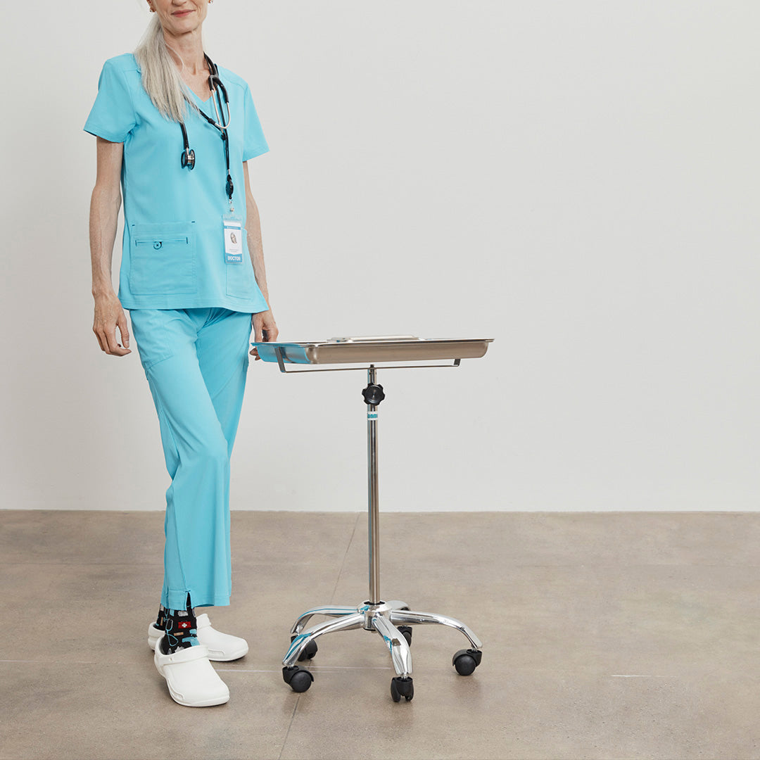 Person in light blue medical scrubs standing next to a height-adjustable table on a plain background