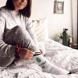 Woman wearing a matching cozy sweatsuit, sitting on a bed, pulling up a pair of ribbed socks with a varsity black double stripe at the top cuff, and a pattern that says "OH KALE NO" and a kale leaf on it. thumbnail