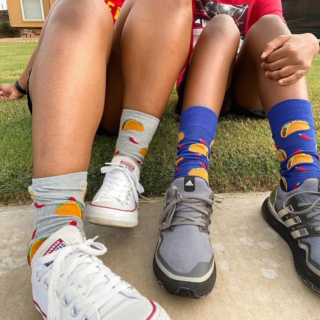 Two pairs of feet wearing colorful taco socks and sneakers on a grassy background