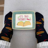 Person wearing taco-themed socks with a decorative plate saying 'It's Taco Tuesday' on a gray surface.