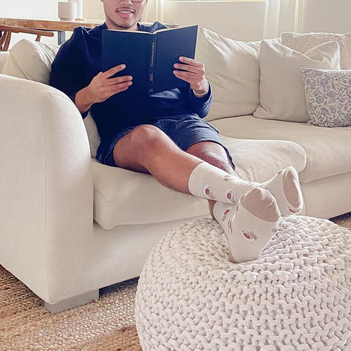Person sitting on a couch reading a book with their feet propped up on a poof wearing HOTSOX coffee cup socks