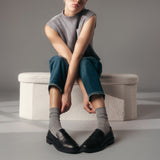 Person sitting on a textured ottoman wearing a sleeveless top, jeans, and black shoes with gray socks. thumbnail