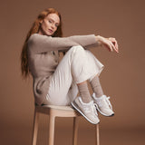 Woman sitting on a stool wearing a beige sweater and socks, white pants, and white sneakers against a brown background