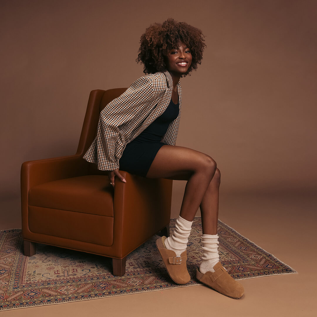 Woman sitting on a brown armchair wearing a checkered shirt, black dress, beige slouch socks and brown shoes on a brown background.