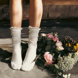 White socks with ruffles worn by a person sitting on a stone surface with flowers. thumbnail