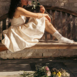 Woman in a white dress and feminine slouch socks sitting on a wooden bench with flowers, surrounded by more flowers on the ground. thumbnail